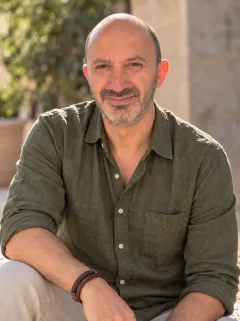 Raul Ortegon-Bolio, smiling, in an olive-green shirt with rolled-up sleeves, sitting outdoors in the sun with plants and a stone wall behind him.